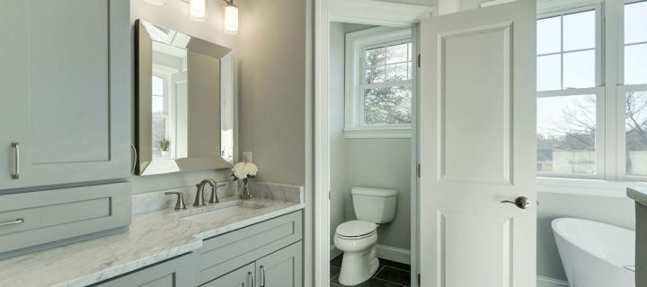 A bathroom remodel with a corner toilet area with a door, a freestanding bathtub, and a vanity area with marble countertop, and a wall-mounted light fixture